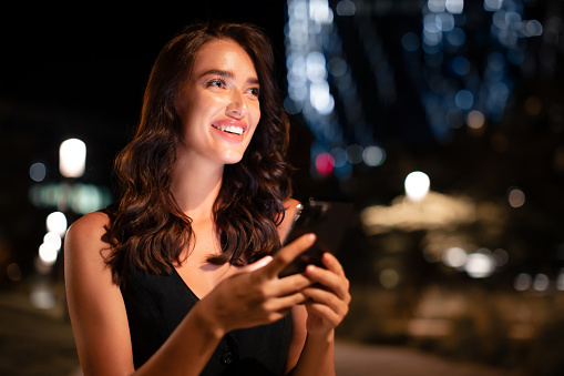 Young smiling caucasian woman on urban city street at night using cellphone and looking aside with bokeh and neon city lights on background, copy space