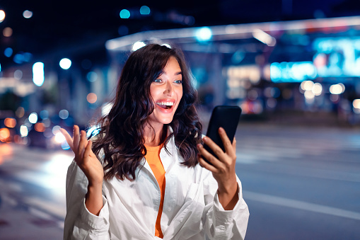 Surprised young woman looking at smartphone screen with excitement, reading message while walking through night city street full of neon lights, copy space