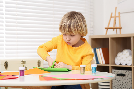 Cute little boy cutting green paper at desk in room. Home workplace