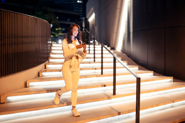 istock Young stylish woman holding cellphone while going down the illuminated stairs, walking in the city at night, full length 1786490761