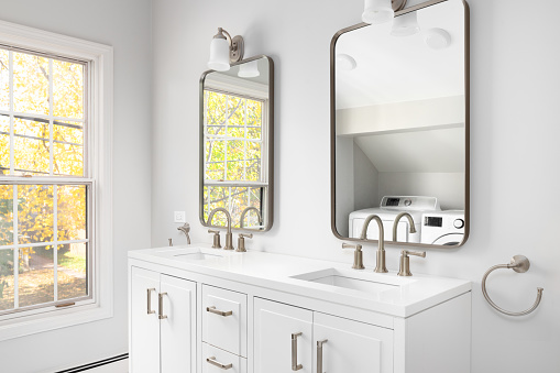 A bathroom detail with a white cabinet, bronze faucets and mirrors, a view of the washer dryer in the reflection, and fall colors outside.