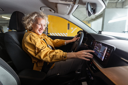 Photo of a smiling woman going to work with her car. She is searching for a good radio station while driving.