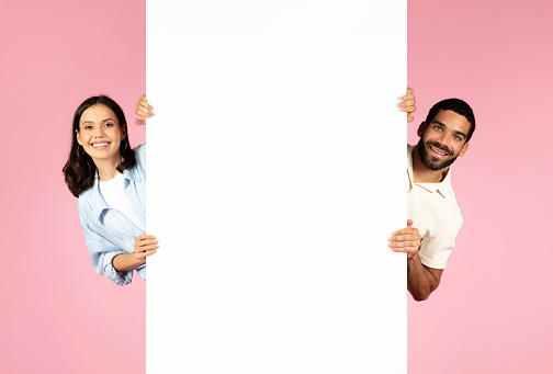 Positive cheerful beautiful millennial hispanic man and woman posing with vertical blank white advertising board, isolated on pink background, blank space for nice offer, deal, text, mockup