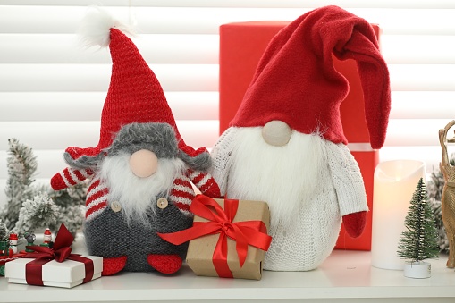 Funny Christmas gnomes and gift boxes on commode in room with festive decoration