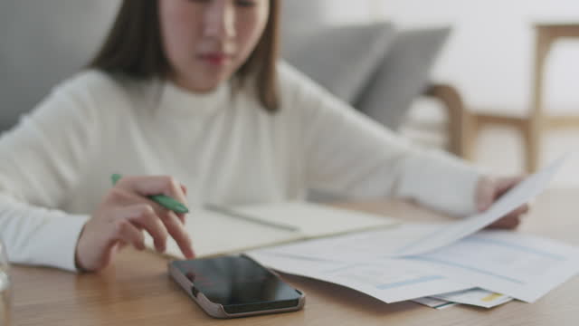 Close-up shot of an Asian woman writing a list of debt in a notebook calculating her expenses with a calculator with many invoices while staying at home.