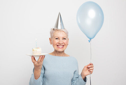People, party, aging and maturity concept. Beauitful middle aged female wearing conical hat celebrating birthday, posing isolated with cupcake with candle and blue balloon in her hands. Happy time.