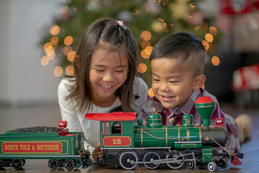 Two young children of Asian decent, take a close look at their new train on Christmas morning.  They are dressed in Christmas outfit and have big smiles on their faces as they enjoy their new toy.