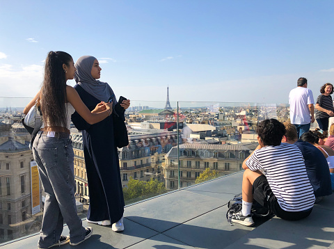 Paris, France: Young Arab women with cameras on the Galeries Lafayette rooftop with the Paris cityscape at their feet and the Eiffel Tower in the distance.