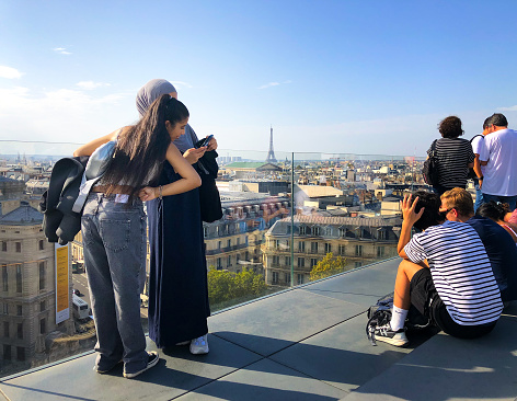 Paris, France: Young Arab women with cameras on the Galeries Lafayette rooftop with the Paris cityscape at their feet and the Eiffel Tower in the distance.
