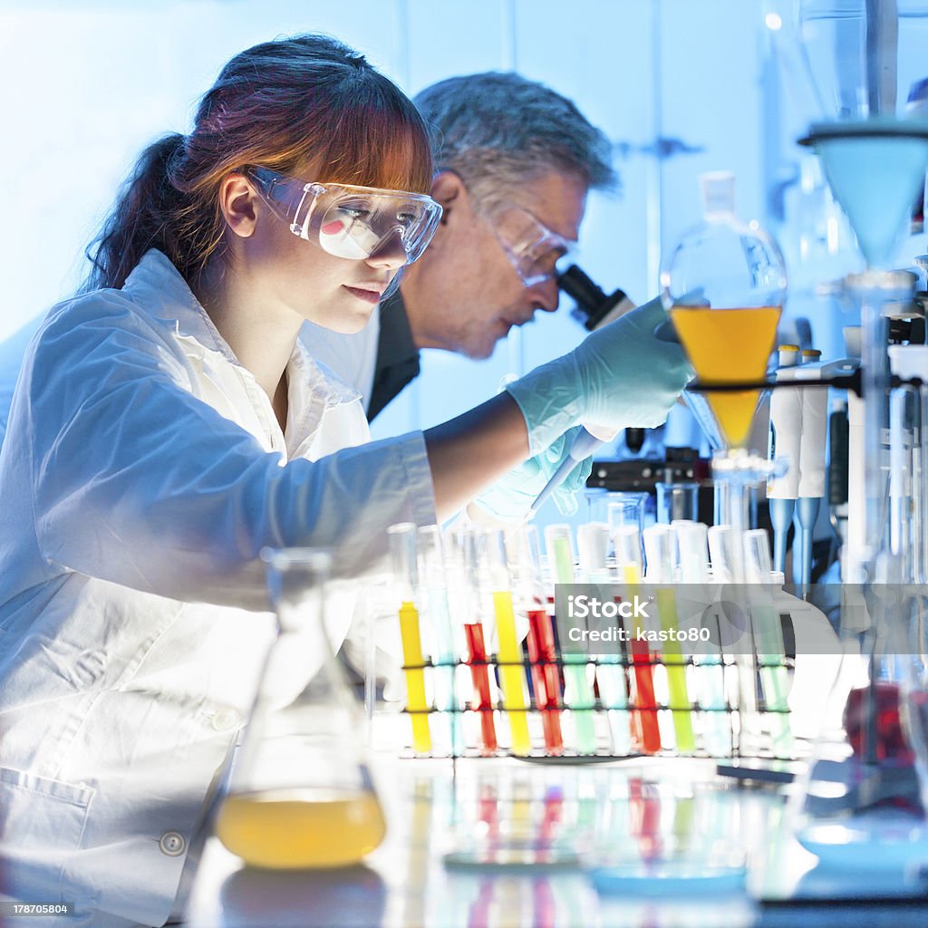 Health care professionals working in laboratory. Attractive young female scientist and her senior male supervisor pipetting and microscoping in the life science research laboratory (biochemistry, genetics, forensics, microbiology..) Adult Stock Photo
