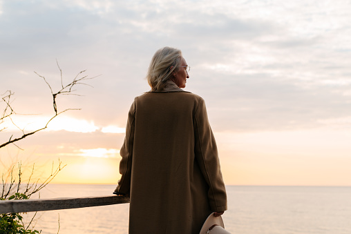 Rear view of senior caucasian woman wearing glasses and coat standing on seashore against sunset sky and looking away.