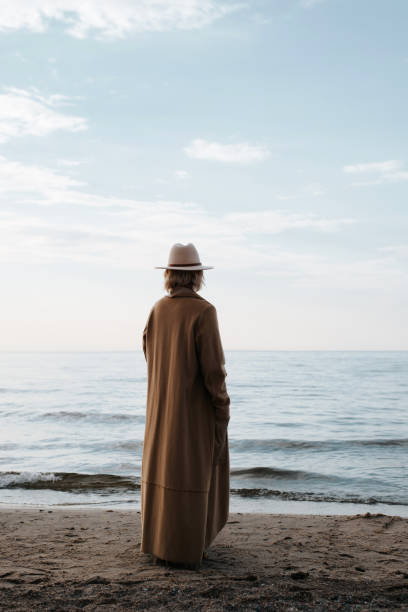 istock Lonely senior woman alone standing on seashore and looking thoughtfully into horizon. Back view of stylish elderly beautiful lady wearing hat, brown coat, mood of loneliness in nature 1787119716