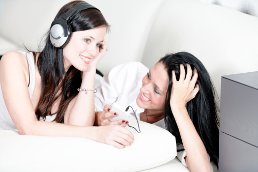 Two beautiful young woman listening to music on headphones at home on the sofa.