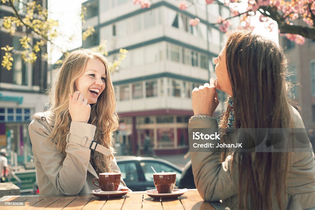 Two women laughing together while drinking coffee at a cafe Two young women talk and drink coffee in cafe, outdoors Girlfriend Stock Photo