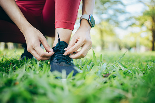 A young woman runner is outside in the morning, preparing for a jog. She is seen bending down, tying her shoelaces, ensuring her running shoes are securely fastened before she begins her run