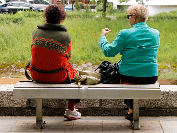 Two female friends resting on bench in park and communicate.