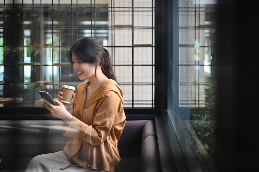 Attractive young businesswoman with takeaway coffee using mobile phone at corporate office break out area.