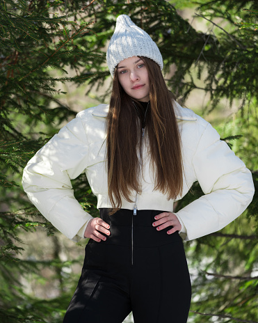 Portrait of beautiful brunette teenage girl arms akimbo, looking at camera. Fashion model standing in winter pine forest. Waist up teen female in hat, white puffer bomber jacket, black pants. Part of series of photographs