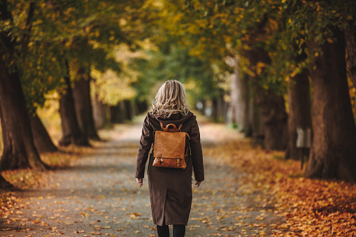 Stylish woman with brown coat and leather backpack walks in alley at autumn park. Beauty and fashion lifestyle