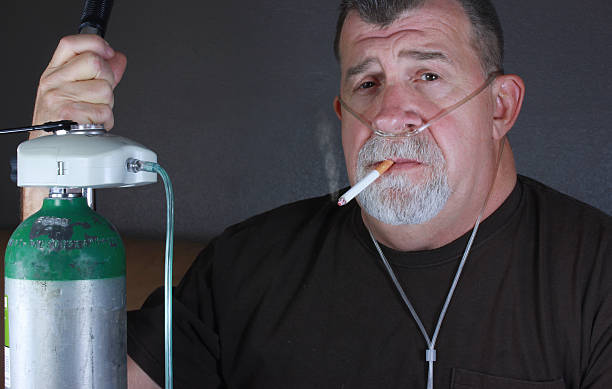 Adult Man Smoking with Cannula and Oxygen Tank A adult man dangerously smokes a cigarette while wearing an oxygen cannula while holding on to an oxygen gas tank. tracheotomy tubing stock pictures, royalty-free photos & images