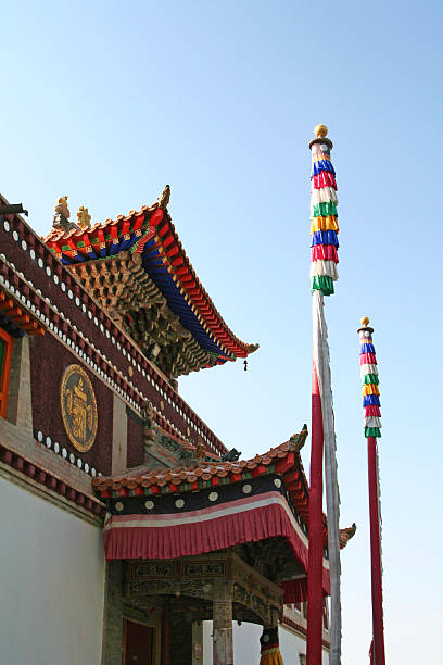 The intricately decorated golden and red roof of a traditional Thai temple is majestically framed by vibrant pink bougainvillea flowers and lush green foliage on either side. A serene image of Buddha is prominently featured on the temple's gable, adding to its spiritual allure. Above, a clear, bright blue sky provides a striking backdrop. The low-angle shot emphasizes the grandeur and ornate details of the sacred architecture, beautifully contrasted with the natural beauty of the surrounding flora under sunny daylight