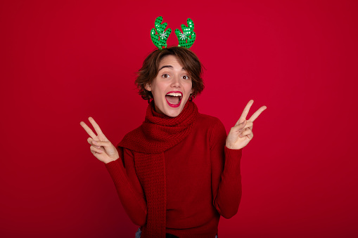 Beautiful young girl in Christmas reindeer horns and scarf is posing and enjoying on party on red background. Merry christmas. Happy new year. Holidays and celebration in winter