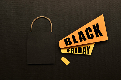 Black shopping bag, yellow and orange colored papers with announcement effect and Black Friday text on it on black background.