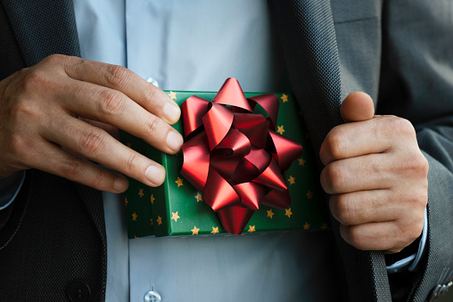 Chest view of an unrecognizable businessman wearing a suit who is about to give a gift box out of his jacket. The green gift box is wrapped with red ribbon.