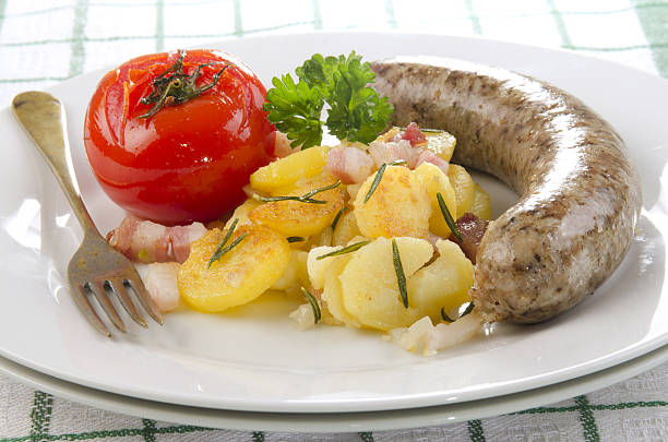 A white ceramic plate with grilled sausages, cherry tomatoes, fried potatoes and herbs on a wooden table, next to it is a gravy boat, a fork and a knife. Close-up.
