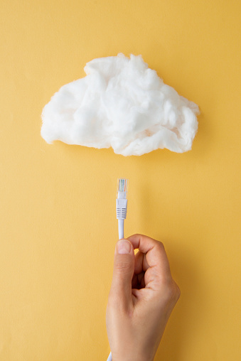 Hand is holding ethernet cable under white cloud on yellow background. Representing internet connection and cloud technology.