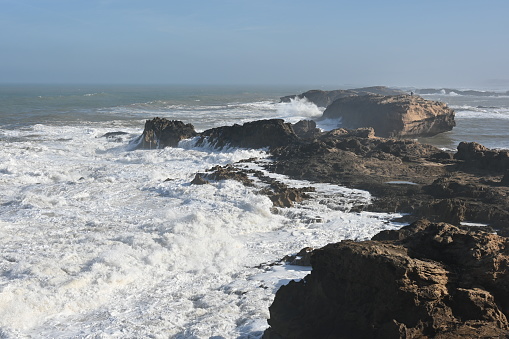 Surf on the Atlantic coast near Essaouira in Morocco, foamy water, rocks and gray-blue sky.