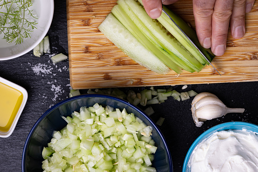 A chef is cutting cucumbers on a chopping board, surrounded by ingredients for the Bulgarian traditional salad made with yogurt, dill and diced cucumbers, known as Snejanka or Milk Salad.