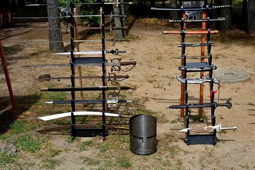 A view of a vast collection of decorative and historic swords, knives, armour, targes, shields, and other medieval military equipment shown on wooden racks and cloth covered tables seen in summer