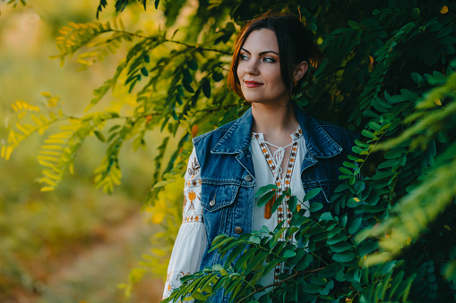 Smiling attractive hippy woman on nature background. Young lady in white embroidery shirt, denim waistcoat. Summer fashion, hipster, ethno, folk lifestyle.