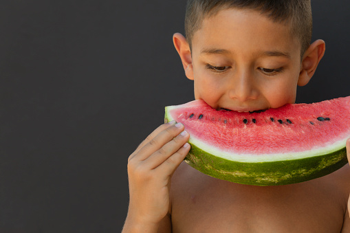 Little boy  eating a slice of watermelon in front of black background with a cute smile.