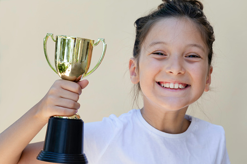 Little girl is holding golden trophy award with a toothy smile and is looking at camera in front of white background. Representing winner and first price of competition.