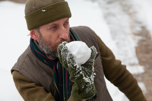 Snow games in the air. A man with a beard in a warm vest and a hat throws snowballs in the yard of a private house on a frosty day