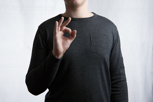 Young man shows OK sign in gray sweater on light gray background. A sign of positivity, high class.