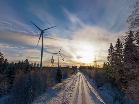 Scenic aerial photo over forest winter road with windmills standing in row at the left side in forest. Road in the middle of view. Large wind turbines with blades