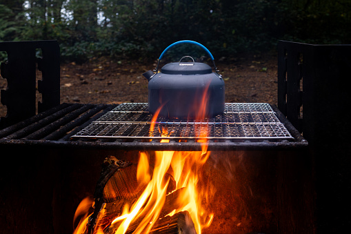 A tea kettle boils water as flames from a camp fire warms the water