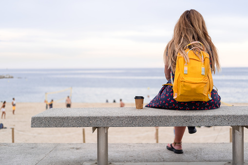 unrecognizable curvy blonde mature woman sitting on a bench in front of the ocean watching a summer beach volleyball game. she is having a coffee to go.