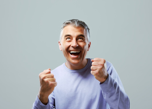 Portrait of excited mature man, raising his fists on grey background.
