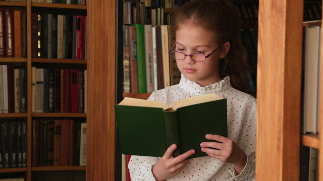 Kid girl in glasses reading book in old public library. Child in bookstore
