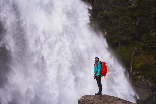 one mature adult woman with backpack standing on rock close to huge waterfall in the mountains