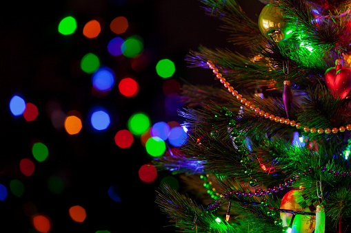 A green Christmas tree festively decorated with toys and luminous garlands stands in a dark room close-up. In the background, colorful bokeh lights