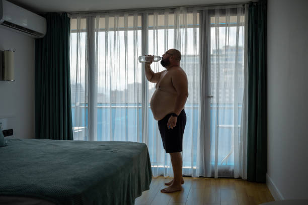 uomo assetato in sovrappeso che sente disidratazione bevendo acqua in camera d'albergo quando fa caldo. - men naked water male foto e immagini stock