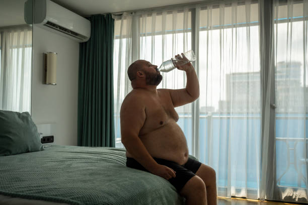 uomo in sovrappeso che beve acqua seduto a letto in camera d'albergo in vacanza nella stagione calda estiva. - men naked water male foto e immagini stock