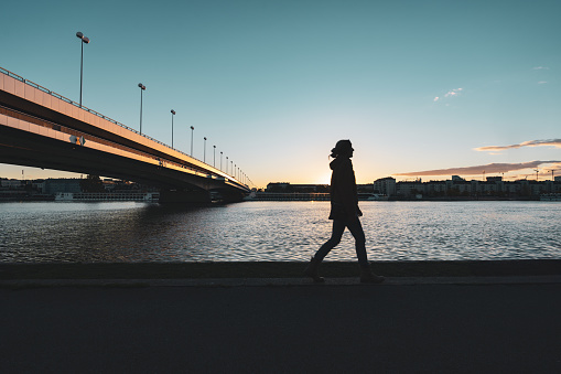 one woman walking alone along danube river in twilight on cold autumn evening, bridge over water, person in silhouette