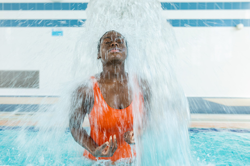 Black young woman massaging her back while closing her eyes under a water jet at the heated swimming pool during a winter day