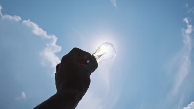 Male Hand Holds Light Bulb Against the Blue Sky and the Bright Sun